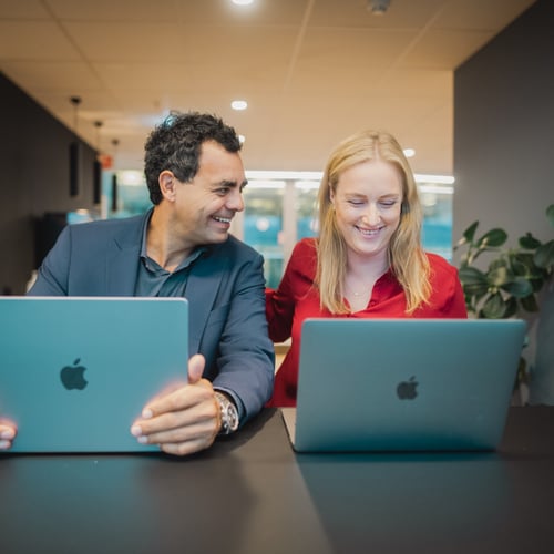 To kollegaer sitter sammen og smiler foran hver sin PC