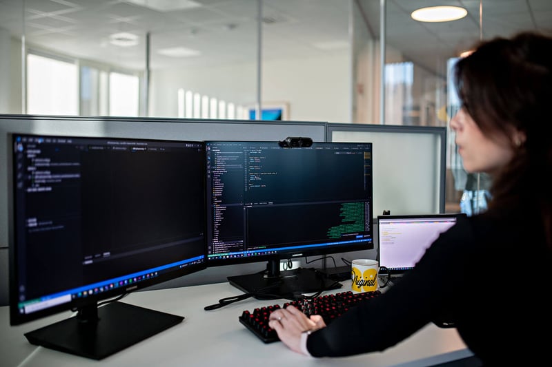 Utvikler – ved pulten – skriver kode på sine to skjermer med hennes gule kopp lett tilgjengelig